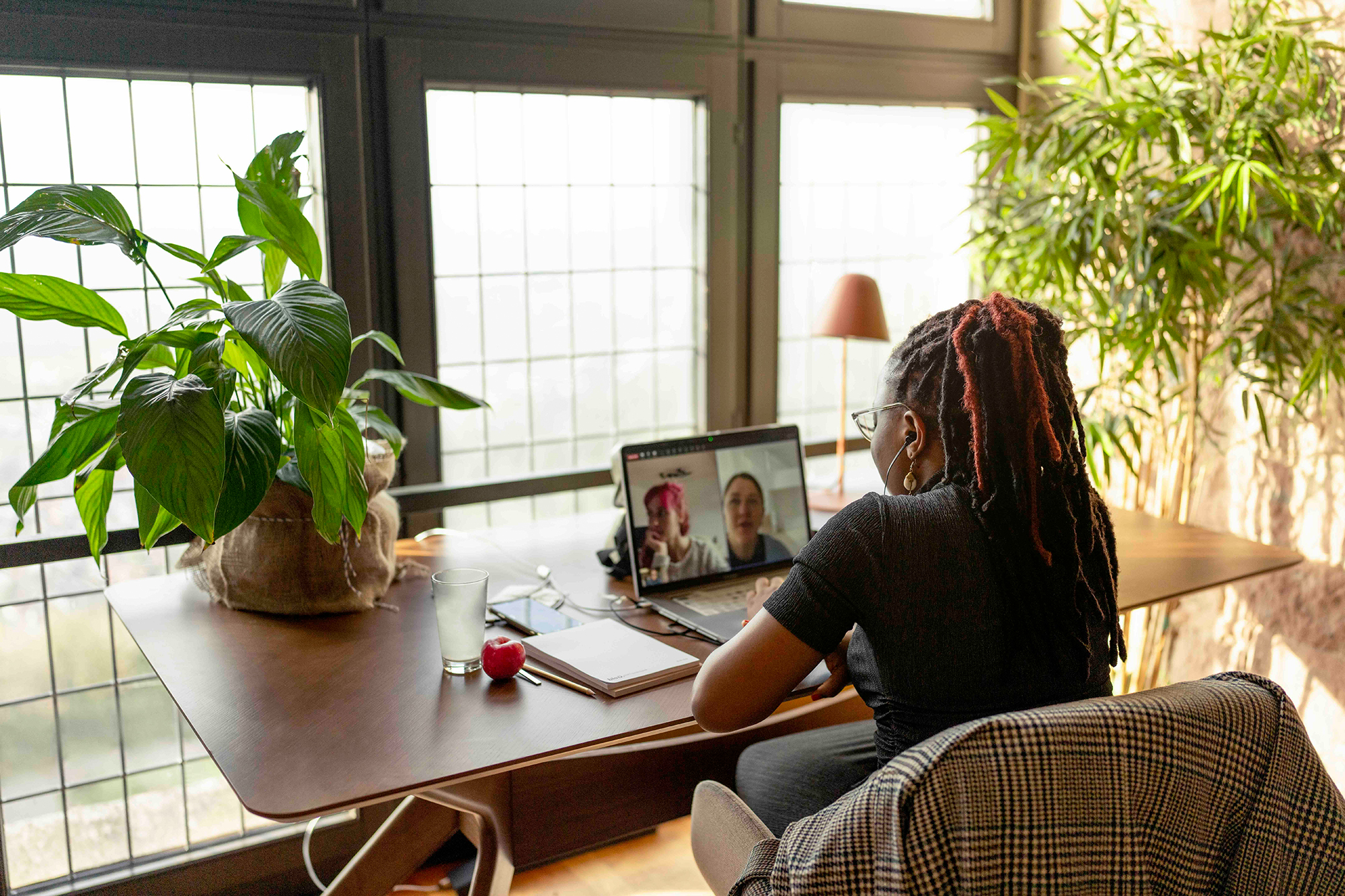 Woman sitting alone at a round table in a room with plants and large windows.