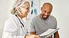 Ältere Ärztin in weißem Kittel spricht mit einem jungen Mann und hält ein Tablet.