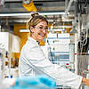 Frau in weißem Kittel lächelt in einem industriellen Labor vor Maschinen.