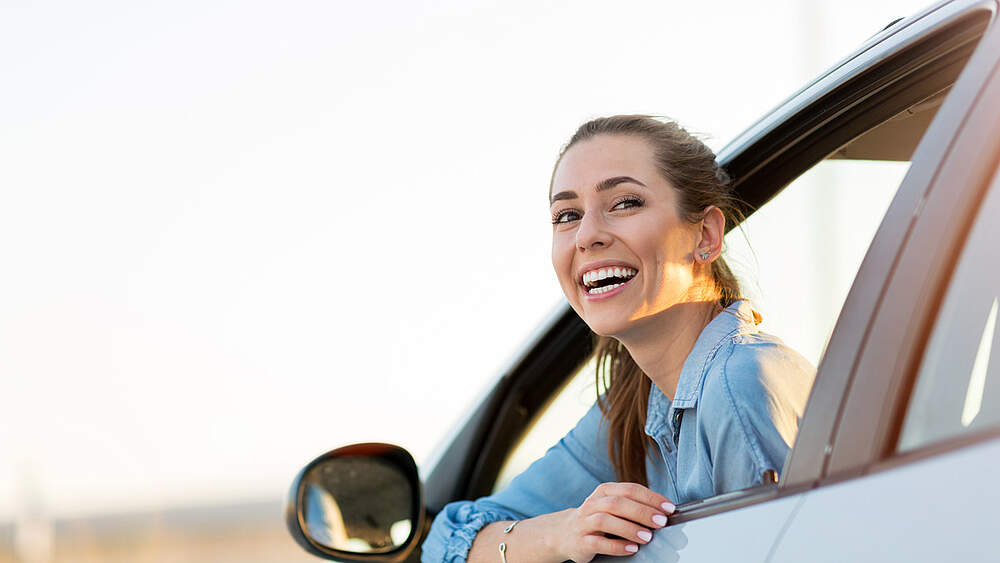 © AdobeStock /Pikselstock A Woman smiles out of the drivers door windows of her car.