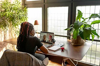 Frau sitzt von hinten an einem runden Tisch in einem lichtdurchfluteten Raum mit Pflanzen.
